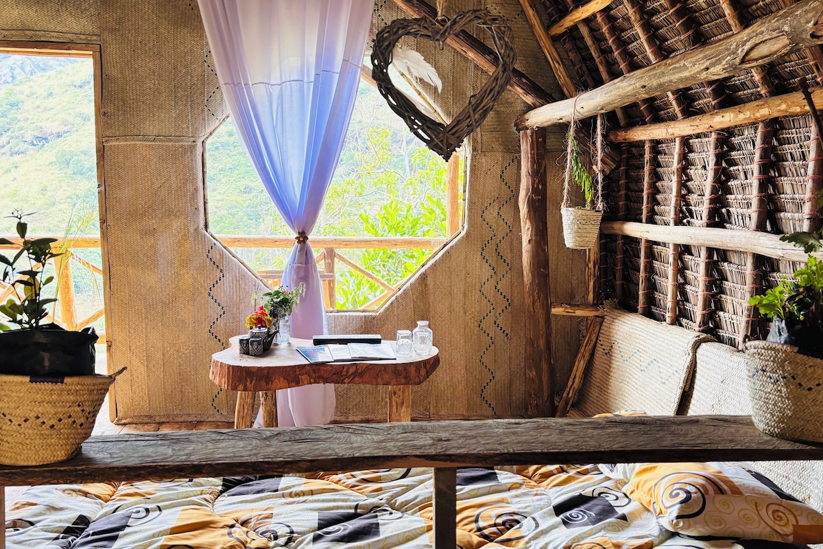 Une pièce rustique avec une grande fenêtre, une petite table sur laquelle est posé un vase, et un panier en osier.