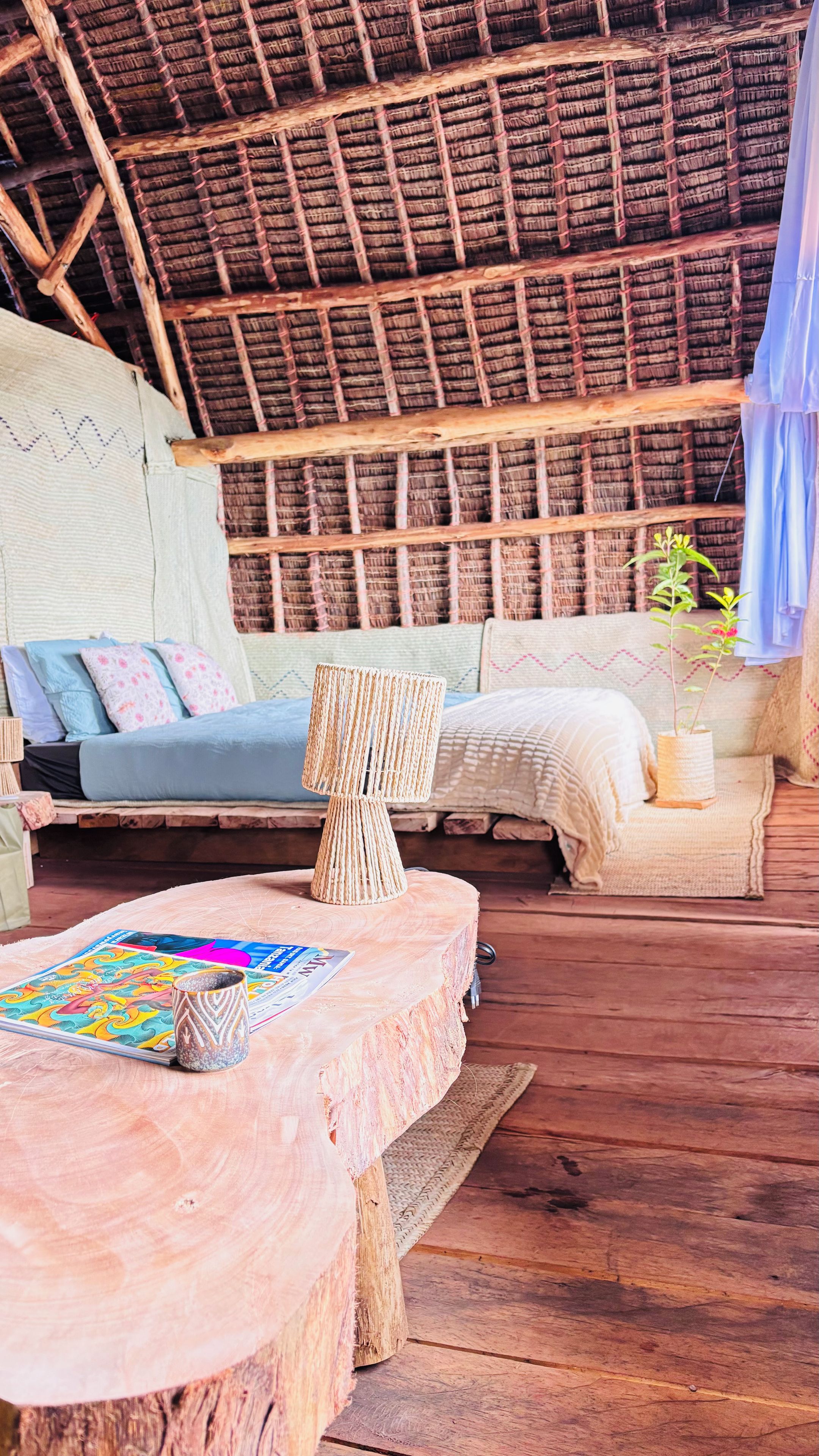 Une pièce rustique avec un toit de chaume, un plancher en bois, un canapé, une lampe en osier et une petite table sur laquelle repose un magazine.