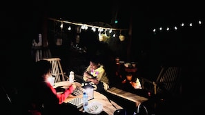 Un espace extérieur à l’éclairage tamisé, aménagé pour les repas, avec des guirlandes lumineuses, une table sur laquelle se trouvent une bouteille et un jeu d’échecs, et un foyer au fond.