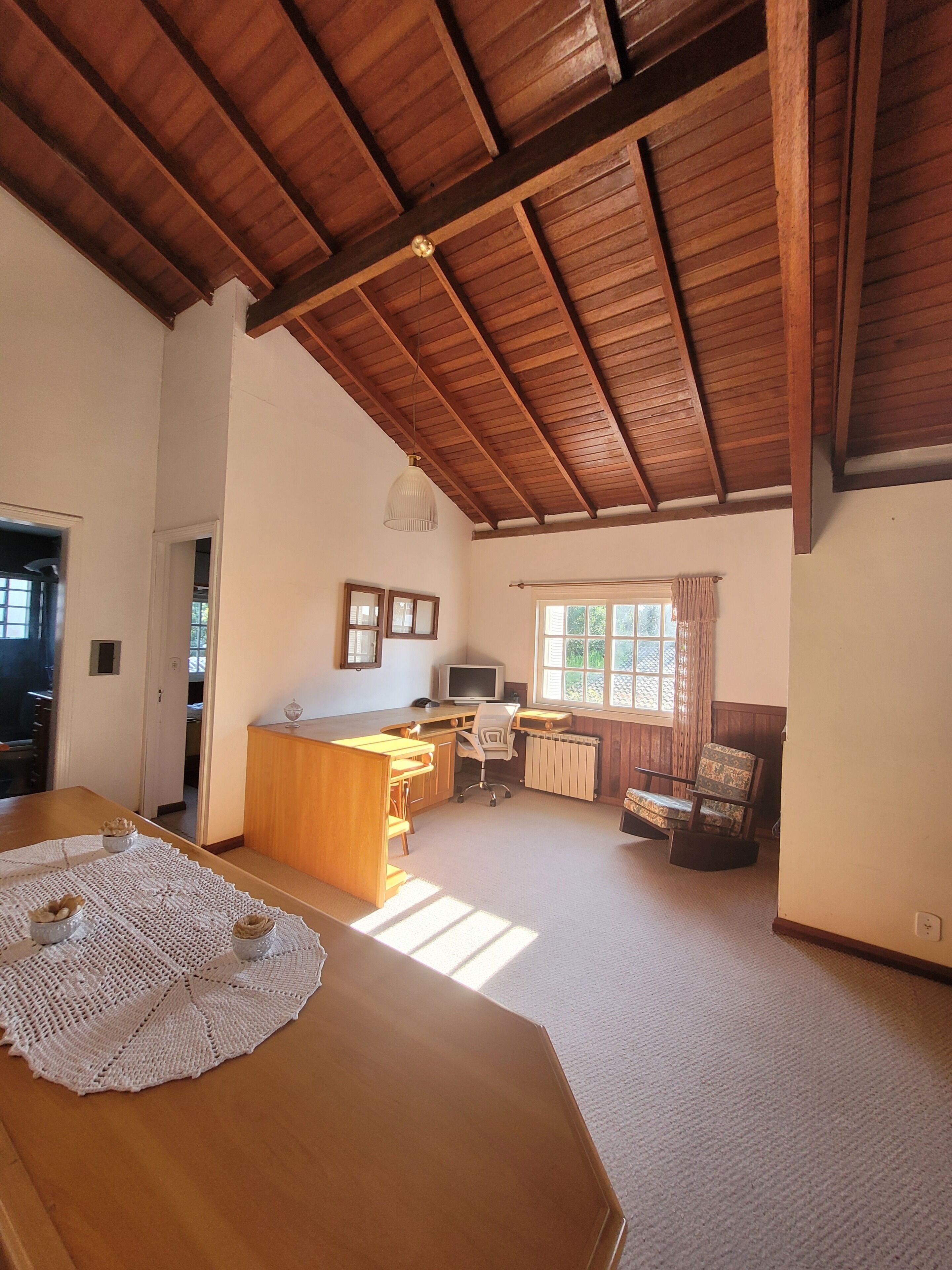 A spacious room with a wooden ceiling, a kitchen area with a dining table, and a window with curtains.