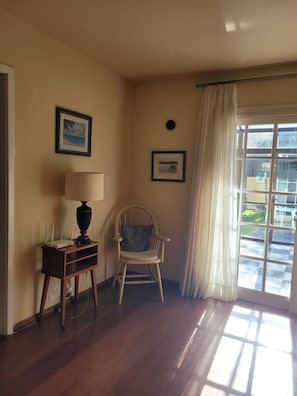 Una habitación con piso de madera, una silla con cojín, una mesa con lámpara y dos cuadros en la pared.