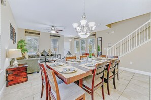 Un coin repas avec une table en bois dressée pour six personnes, entourée de chaises, et un ventilateur de plafond ainsi qu’un lustre au-dessus.