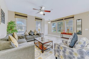 Un salon spacieux doté d’un ventilateur de plafond, de grandes fenêtres et d’un mélange de canapés et de fauteuils.