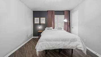 Een slaapkamer met een bed, een nachtkastje, een lamp, een raam met gordijnen en een kunstwerk aan de muur.