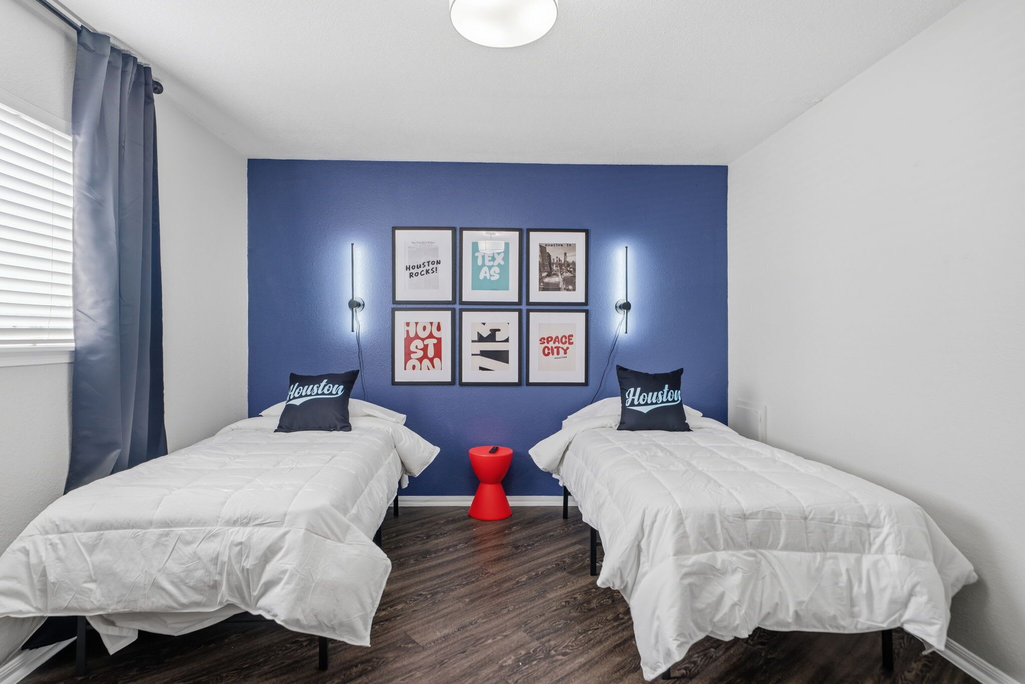 Une chambre avec deux lits, un tabouret rouge et des œuvres d’art murales.