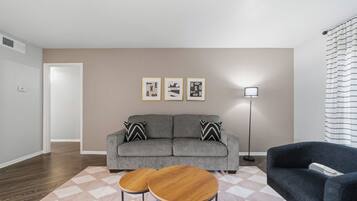 Un salon avec un canapé gris, deux petites tables rondes en bois, une lampe sur pied et trois tableaux encadrés accrochés au mur.