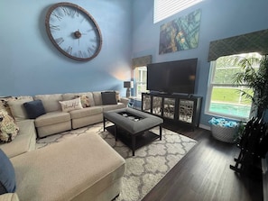 A living room with a sectional sofa, a coffee table, a large wall clock, a flat-screen TV, and a window with a view of a pool. - Peaceful Backyard Paradise (Haines City)