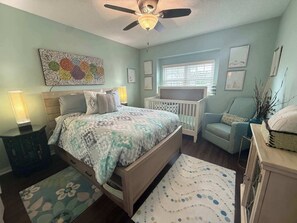 A bedroom with a bed, a crib, a chair, a ceiling fan, and artwork on the walls. - Peaceful Backyard Paradise (Haines City)