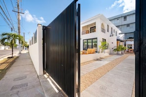 Een omheind woongebied met een modern wit gebouw, een verharde oprit en een klein terras met tuinmeubilair.