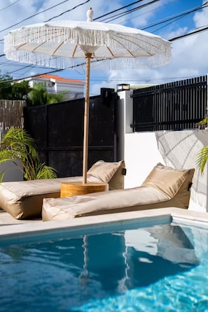 Een zwembadgedeelte met een witte parasol, beige ligbedden en een houten tafel.
