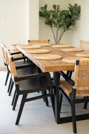Mesa de jantar com jogos americanos trançados, cadeiras pretas com assentos de tecido e uma planta em vaso ao fundo.