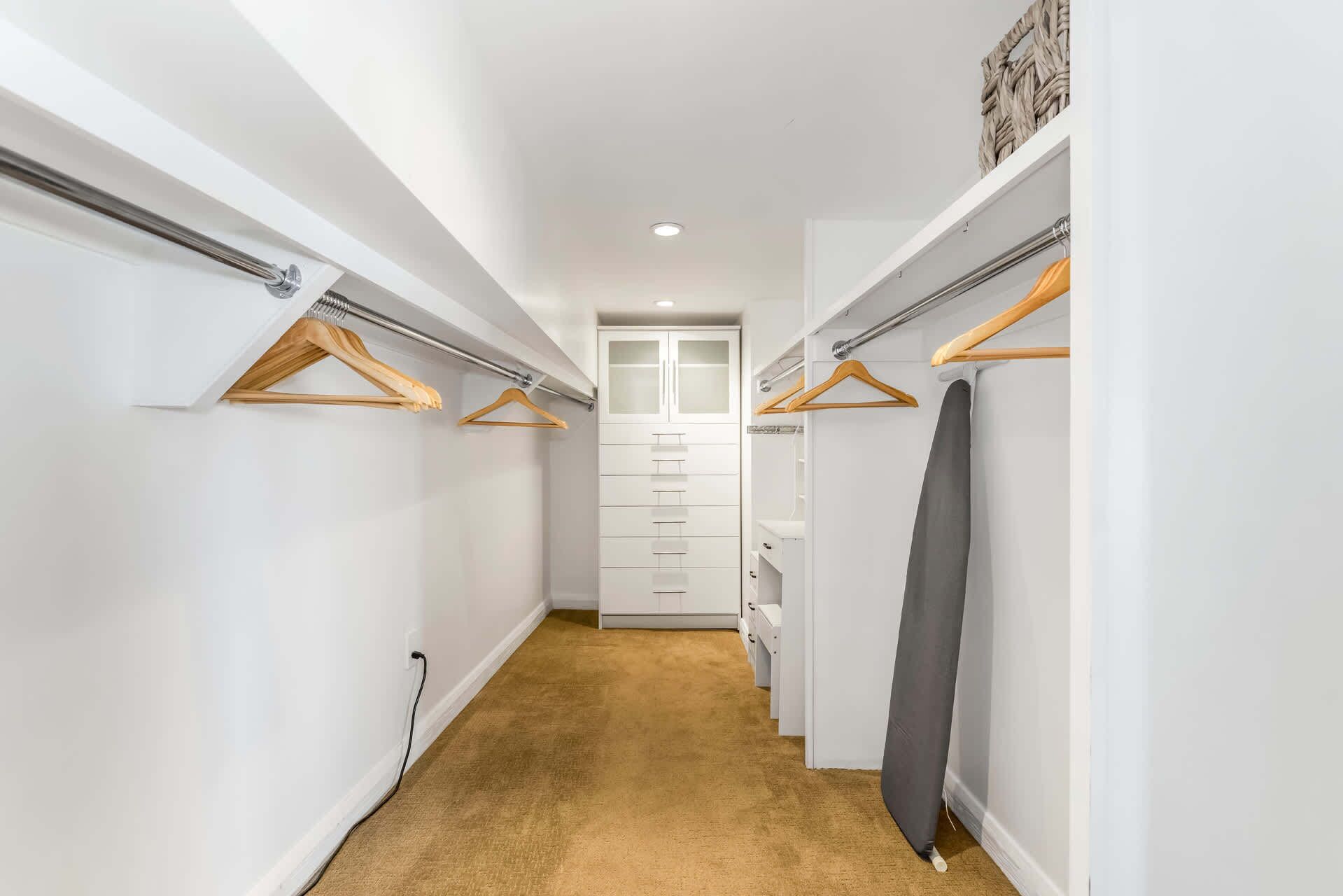A narrow hallway with a carpeted floor, white walls, and a closet with hanging clothes and a large mirror.