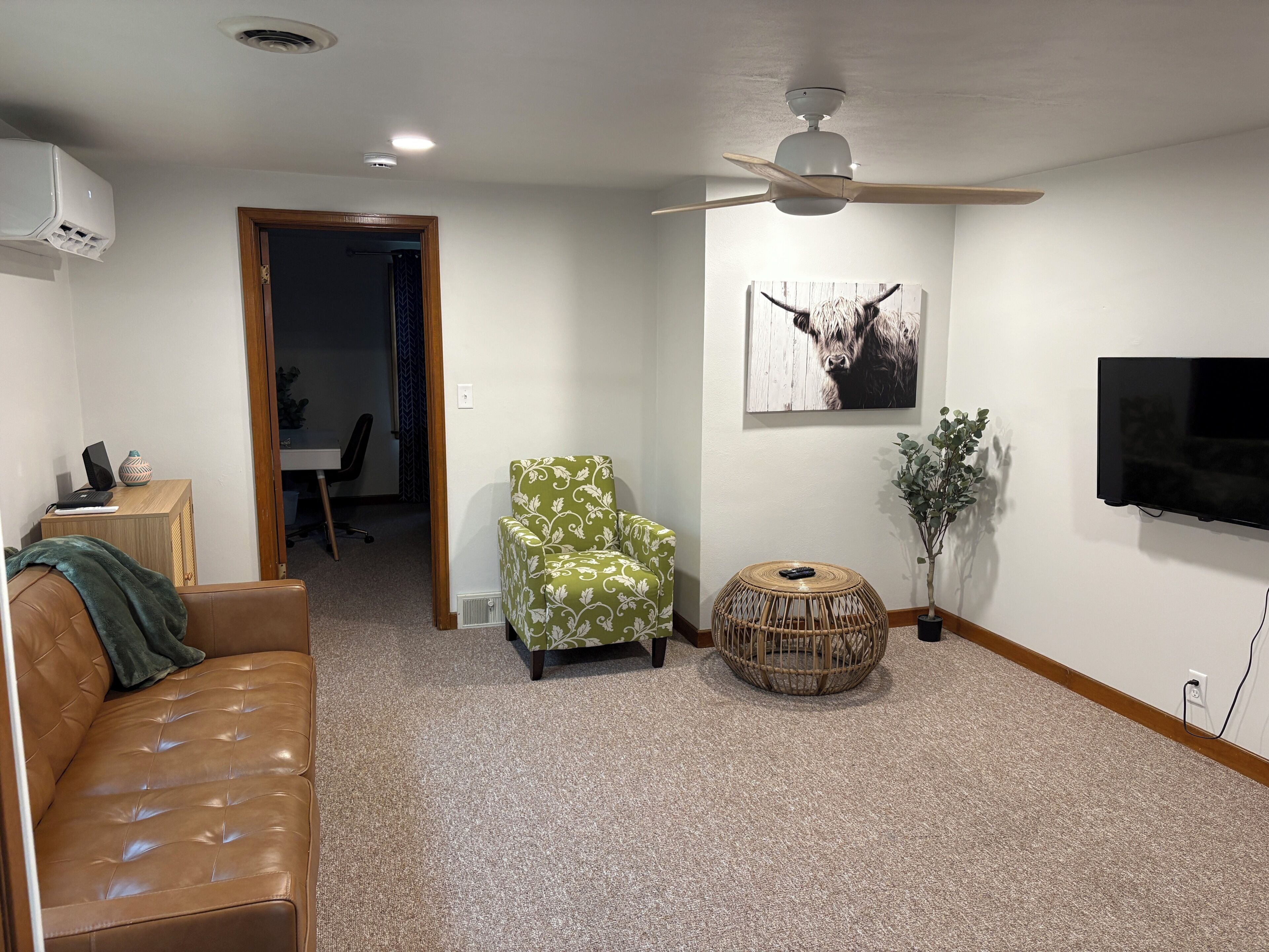 A living room with a brown leather couch, a patterned armchair, a round wicker side table, a television mounted on the wall, and a ceiling fan.