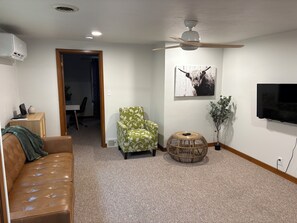 A living room with a brown leather couch, a patterned armchair, a round wicker side table, a television mounted on the wall, and a ceiling fan. - Stay in America's Pumpkin Capital! Furnished Apartment Near Peoria Hospitals (Morton)