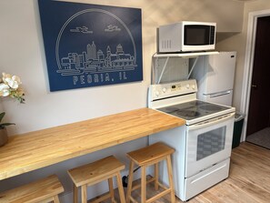 A kitchen with a white stove, microwave, and refrigerator. There is a wooden countertop and two stools. A cityscape artwork is on the wall. - Stay in America's Pumpkin Capital! Furnished Apartment Near Peoria Hospitals (Morton)