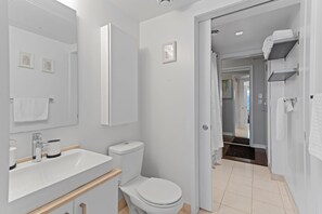 Et moderne badeværelse med hvid vask, toilet og spejl. Et håndklædetørrestativ og en bruseniche er også synlige.