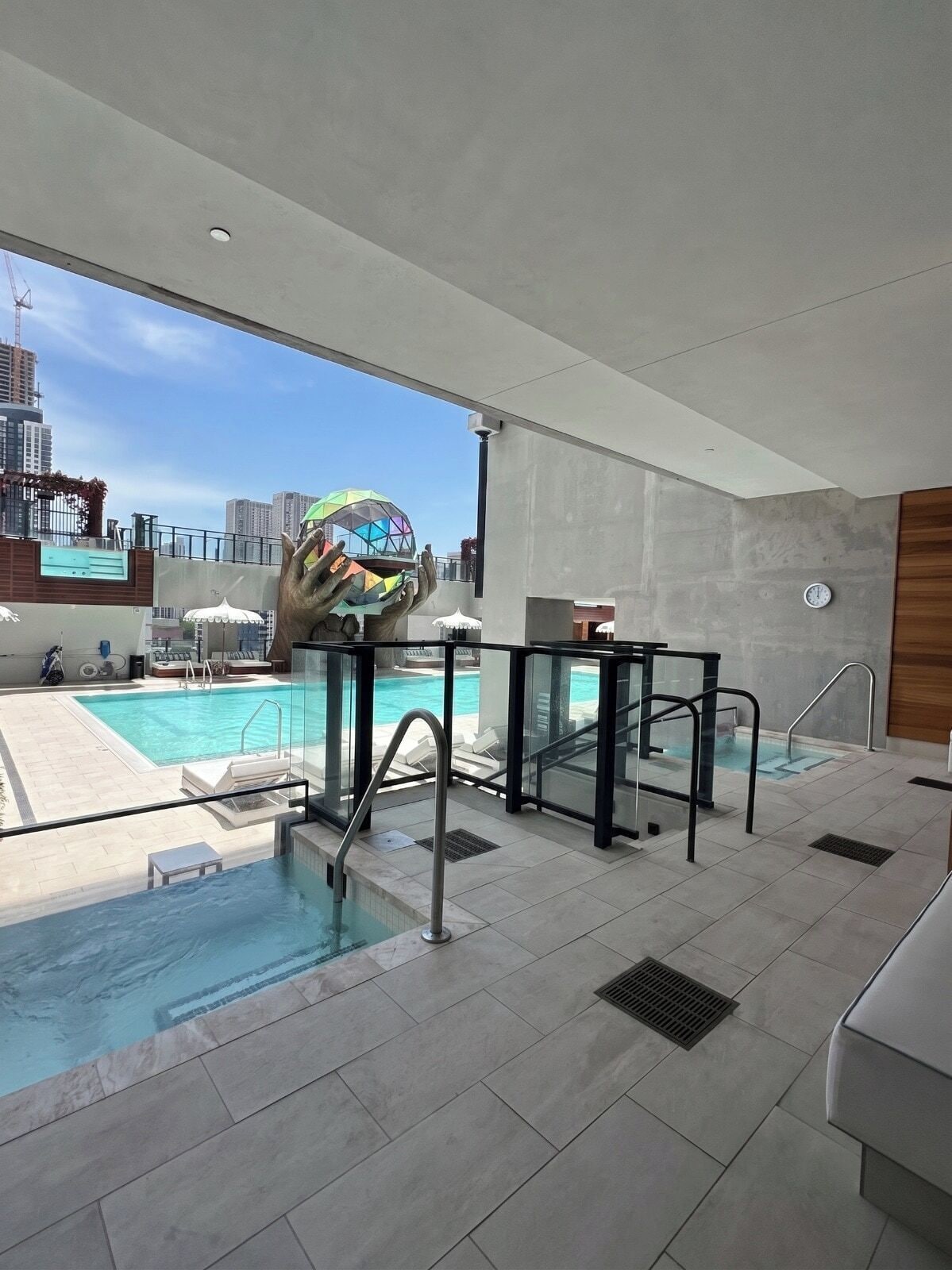 A modern indoor swimming pool with clear glass barriers, a view of the cityscape, and a unique spherical sculpture.