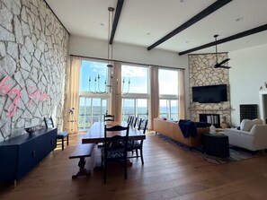 A spacious living room with a dining area, large windows, a stone wall, and a flat-screen TV. - Secluded 4-bedroom house with amazing view of hill country and the Blanco river (Wimberley)
