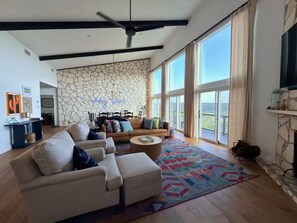 A spacious living room with a stone wall, large windows, a ceiling fan, and a fireplace. - Secluded 4-bedroom house with amazing view of hill country and the Blanco river (Wimberley)