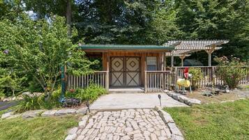 Un bâtiment rustique en bois, doté d’un toit végétalisé, d’un chemin de pierre menant à l’entrée et d’un jardin clos où poussent diverses plantes et fleurs.