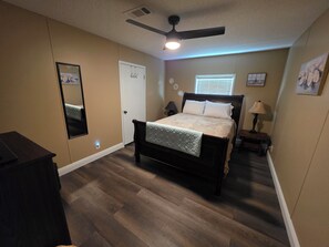 A bedroom with a bed, a ceiling fan, a window with blinds, a bedside table with a lamp, and a dresser. - 1800 sq ft, 3-bed, 2- bath, 2 plus acres in Gulf Hammock Florida. (Morriston)