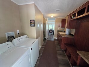 A laundry area with two white front-loading washers, a wooden countertop with a sink, and a hallway leading to a dining area. - 1800 sq ft, 3-bed, 2- bath, 2 plus acres in Gulf Hammock Florida. (Morriston)