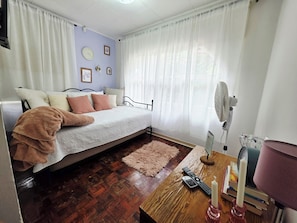 A bedroom with a bed, a wooden table, a fan, and a window with curtains.