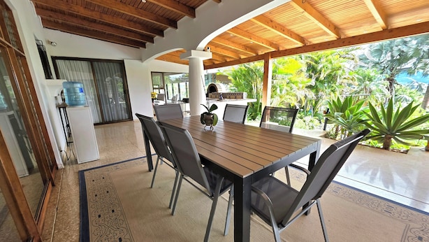 A covered outdoor dining area with a wooden table and chairs, surrounded by tropical plants.