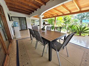 Un espace extérieur abrité, aménagé pour prendre les repas, avec une table et des chaises en bois, entouré de plantes tropicales.