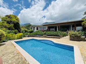 Une piscine bordée de pierres, entourée d’une végétation luxuriante et adossée à un bâtiment doté d’un patio couvert.