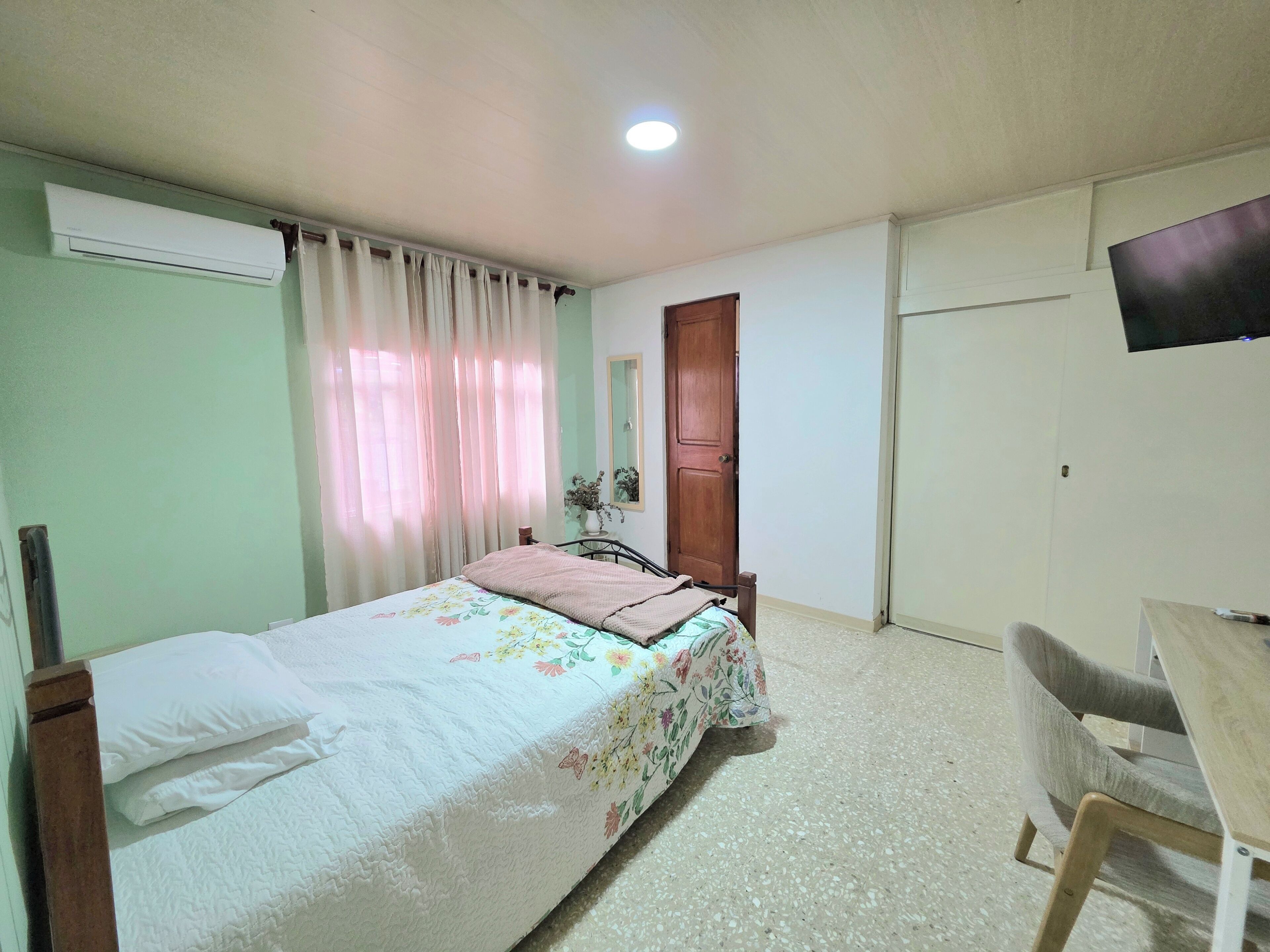 Une chambre à coucher comprenant un lit, un bureau, une chaise, un appareil de climatisation, une porte et un téléviseur.