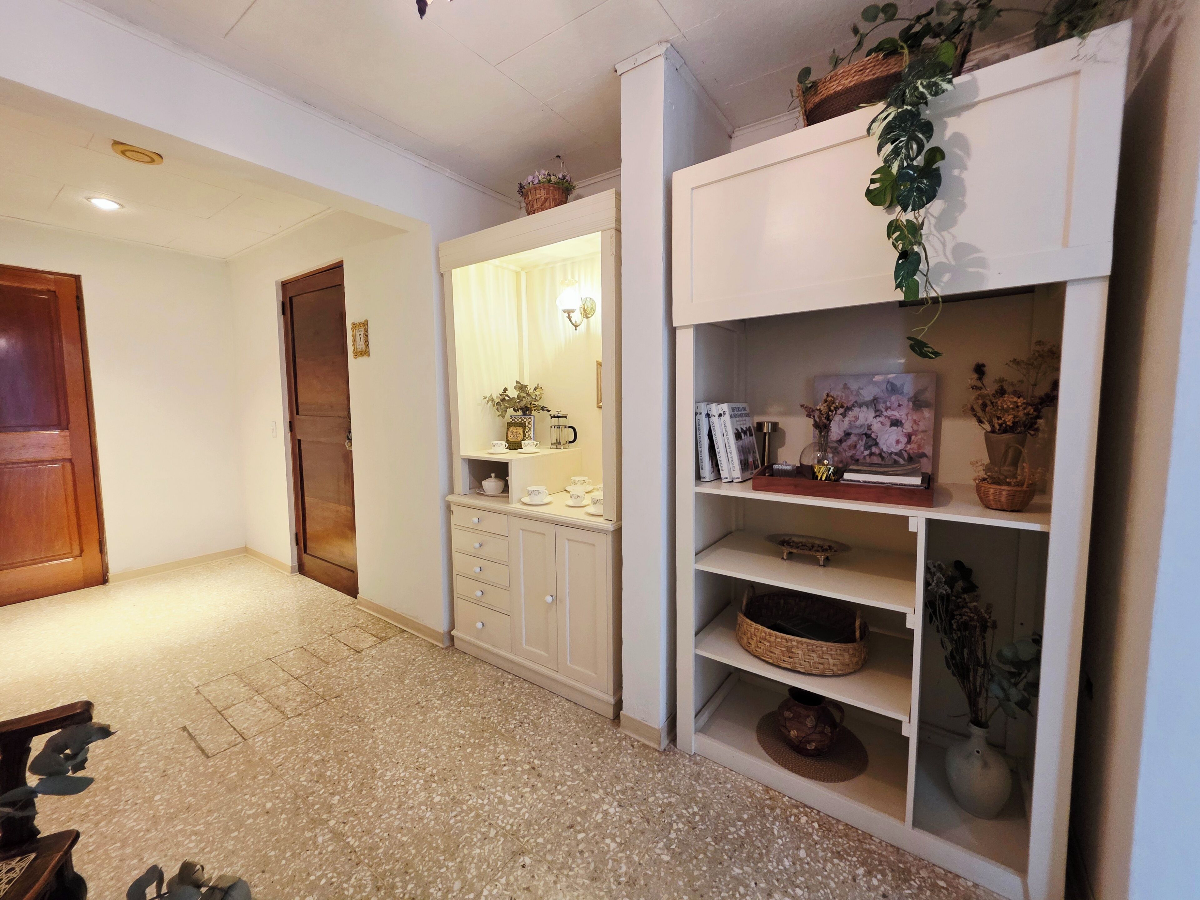 Un pasillo con una puerta de madera, un armario blanco con cajones y un estante con adornos.