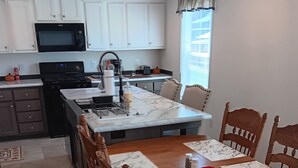 A dining area with a wooden table and chairs, a kitchen in the background with white cabinets and a microwave, and a window with blinds. - 3-bedroom house in peaceful Stacyville  short distance to mt katahdin (Stacyville)