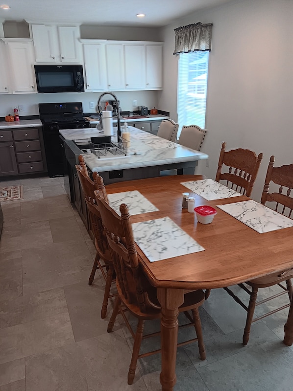 A dining area with a wooden table and chairs, a kitchen in the background with white cabinets and a microwave, and a window with blinds.