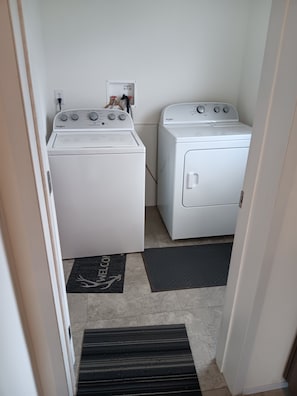Ein Waschraum mit zwei weißen Whirlpool Waschmaschinen und Trocknern, einer schwarzen Fußmatte mit 'WELCOME' und einem Hirschmotiv sowie einer schwarzen Matte auf dem Boden.