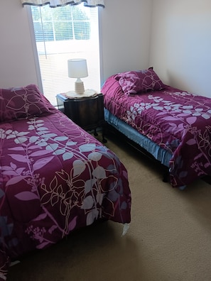 A room with two beds, each with a floral-patterned bedspread, a bedside table with a lamp, and a window with blinds.