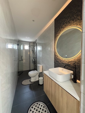 A modern bathroom with a glass shower enclosure, a white toilet, a sink with a black faucet, and a large mirror with a circular light. - Villa with Office, Gym, Jacuzzi & Dryer near CIQ (Johor Bahru)