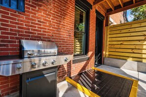 A barbecue grill with four burners, a stainless steel oven, and a built-in sink, set against a brick wall with a wooden deck and a yellow safety barrier. - Louisville Lore | Hot Tub • NuLu • Walk Everywhere (Louisville)