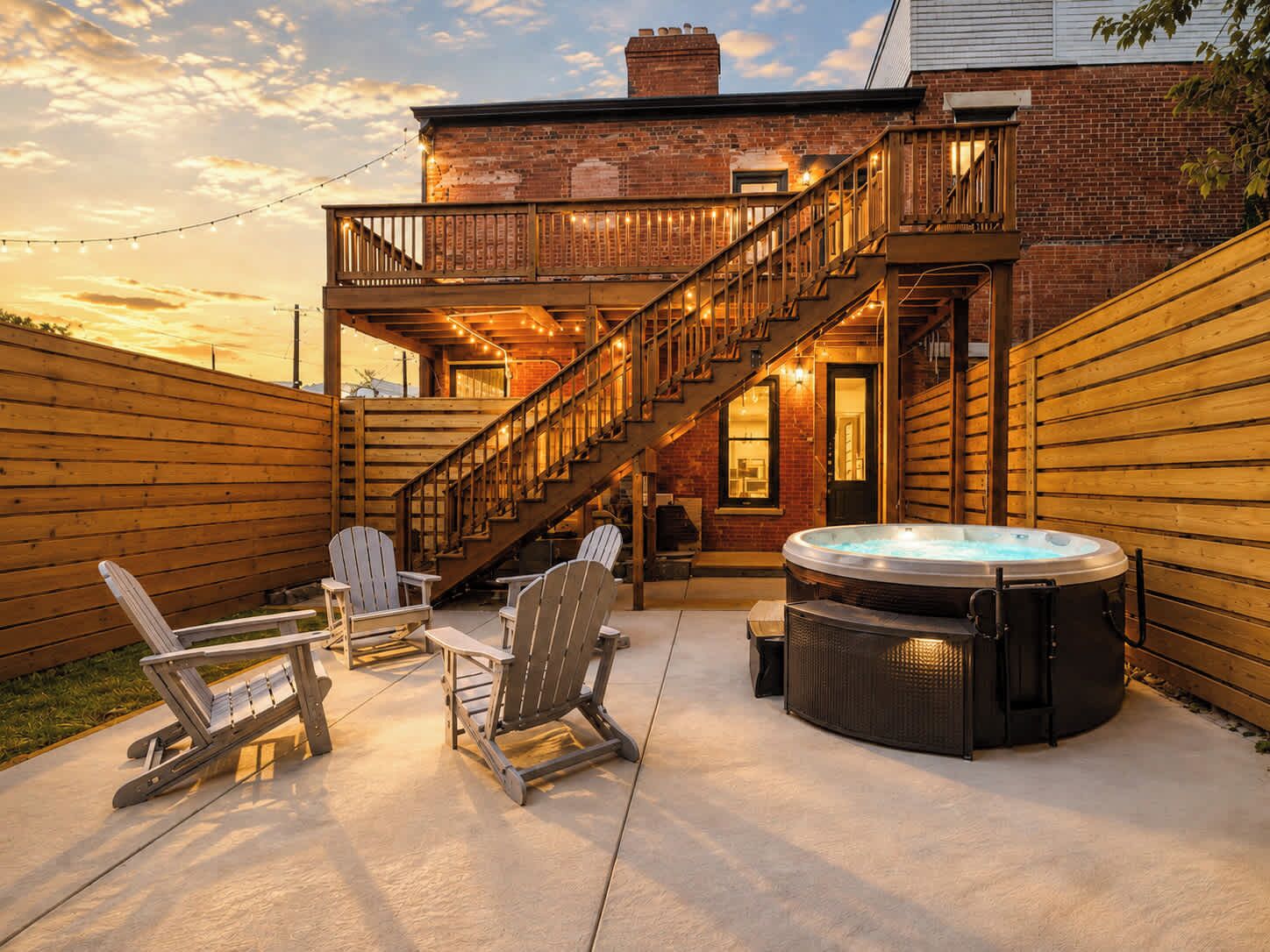 Un jardin avec un jacuzzi, une terrasse en bois, des fauteuils Adirondack et un escalier menant à un balcon situé à l’étage.
