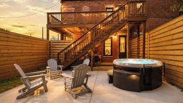 A backyard with a hot tub, wooden deck, Adirondack chairs, and a staircase leading to a second-story balcony.