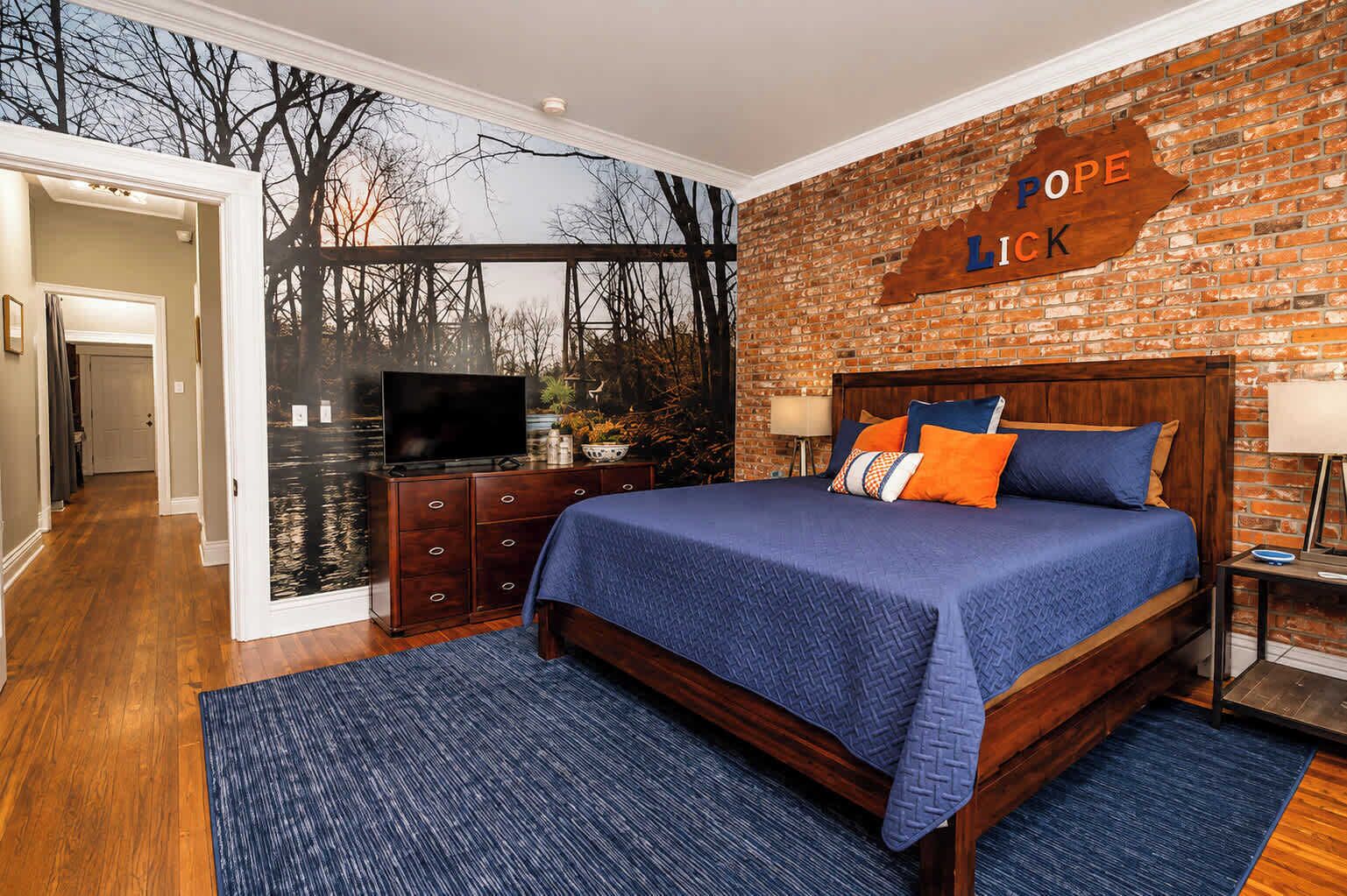A bedroom with a brick wall, a bed with blue bedding, a television on a dresser, and a sign that reads 'POPE LICK'.