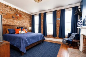 A bedroom with a brick wall, a bed with blue bedding, a chair, and a fireplace. - Louisville Lore | Hot Tub • NuLu • Walk Everywhere (Louisville)