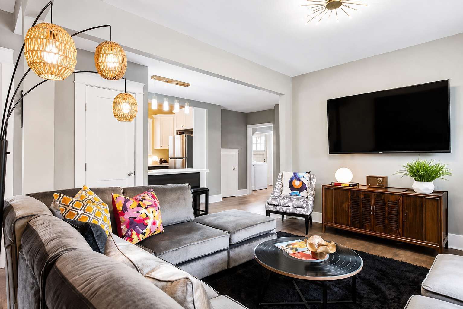 A modern living room with a grey sofa, colorful pillows, a coffee table with vinyl records, a wooden cabinet, and a flat-screen TV.