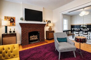 A living room with a brick fireplace, a flat-screen TV, a striped armchair, a yellow armchair, and a dining area with white chairs. - Legends of Louisville | Spacious 6BR • Walkable! (Louisville)
