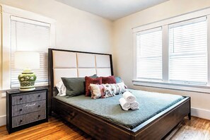 A bedroom with a bed, a bedside table, a lamp, and a window with blinds. - Legends of Louisville | Spacious 6BR • Walkable! (Louisville)