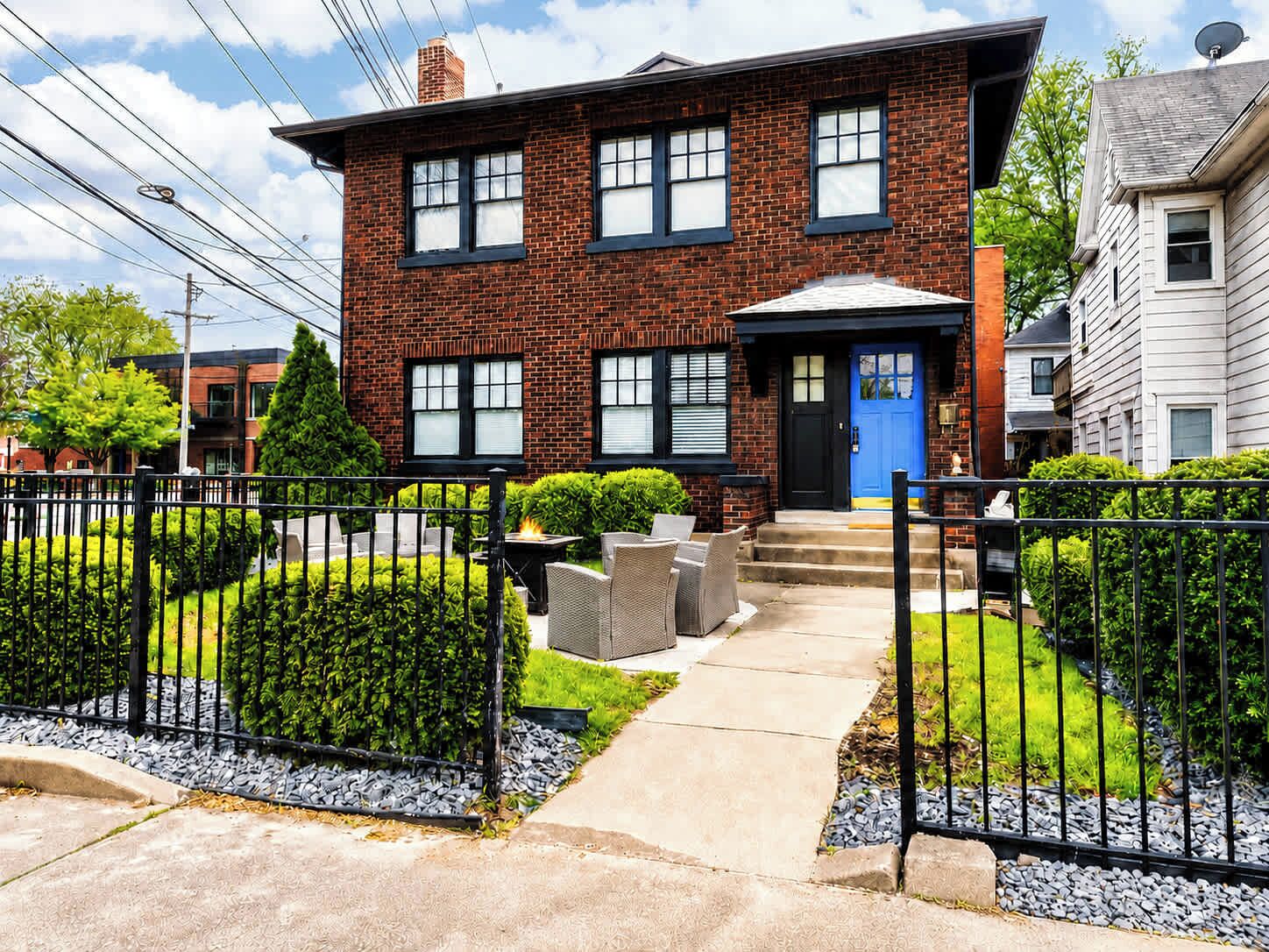 A two-story brick house with a blue front door, a small front porch with outdoor furniture, and a well-maintained garden with hedges and a gravel pathway.