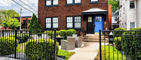 A two-story brick house with a blue front door, a small front porch with outdoor furniture, and a well-maintained garden with hedges and a gravel pathway.