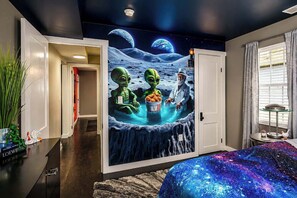 Une chambre décorée d’une fresque murale représentant des extraterrestres sur le thème de l’espace, un lit recouvert d’une couverture à motifs galactiques et une fenêtre avec des rideaux.