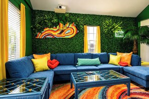 A vibrant living room with a blue sofa, colorful cushions, a glass coffee table, and a green wall with a leafy pattern. - The Wild Side Retreat | Hot Tub • Walk Everywhere! (Louisville)
