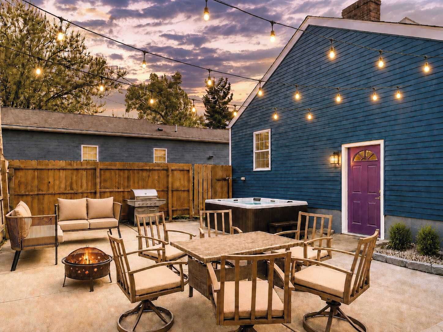 An outdoor patio with a wooden table and chairs, a hot tub, and string lights.